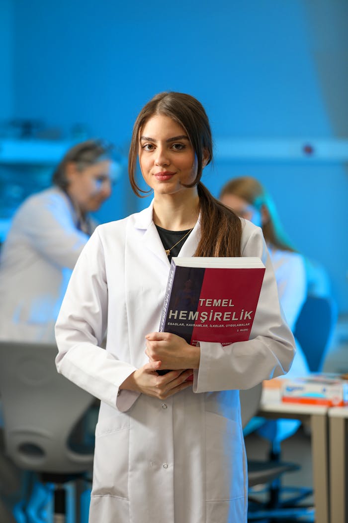 Home Young woman in a lab coat holding a nursing textbook in a classroom setting.
