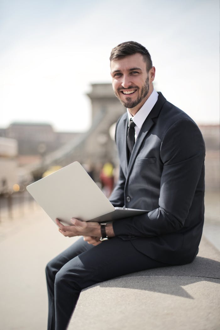 Services Happy businessman in suit holding laptop while sitting on a city bridge, showcasing modern corporate lifestyle.